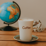The photo shows a cup of coffee on a saucer next to a globe. The cup and saucer are white, and the globe is blue and green. They are standing on a wooden table.