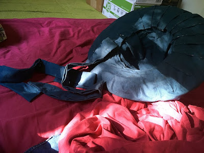 An upside-down straw hat on a mattress with fuchsia sheets, completely lined with pleated medium-blue cotton fabric, pinned with silver pins, and with two long fabric ribbons spilling out of the crown, pinned at intervals. 