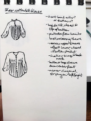 A tiny vertical spiral-bound sketchbook being held in front of a black keyboard. The page is labelled "Sheer cotton/silk blouse" and has two black pen sketches of long-sleeved button-down shirts, both with slightly belled sleeves and thin vertical lines on the lower halves. One has a high band collar and keyhole, and the other a small vee neck and keyhole. On the right side of the page is a column of notes in black pen.