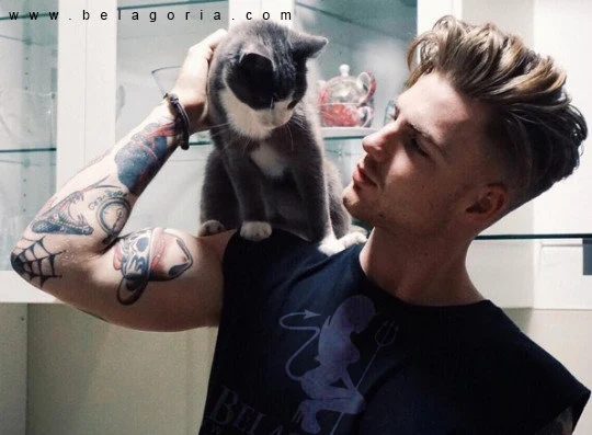 Vemos a un hombre joven con un gato, el chico lleva tatuajes old school en los brazosç