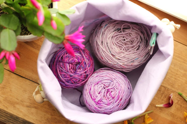 Three shades of purple yarn cakes nestled inside the open project bag with a lavender interior fabric.