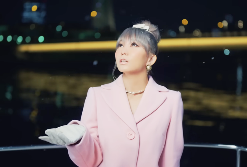 A screenshot from Koda Kumi’s music video for “Tooi Machi no Dokoka de...”. Featuring Kumi stood outside on the boat in a pink coat and white gloves, holding up her hand to catch the snow that’s falling.