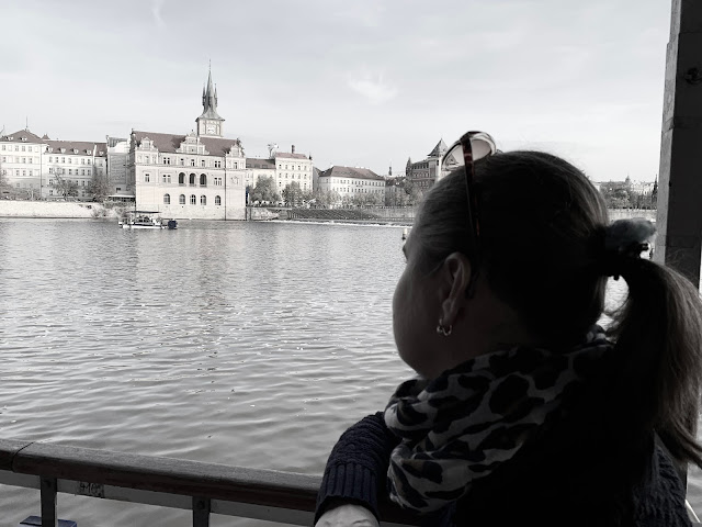 Linda se dívá z paluby lodi na Vltavu. V pozadí je vidět malé plavidlo a stavby historického centra Prahy-