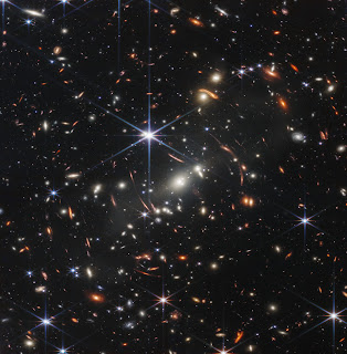 SMACS 0723 (Webb’s First Deep Field) - The background of space is black. Thousands of galaxies appear all across the view. Their shapes and colors vary. Some are various shades of orange, others are white. Most stars appear blue, and are sometimes as large as more distant galaxies that appear next to them. A very bright star is just above and left of center. It has eight bright blue, long diffraction spikes. Between 4 o’clock and 6 o’clock in its spikes are several very bright galaxies. A group of three are in the middle, and two are closer to 4 o’clock. These galaxies are part of the galaxy cluster SMACS 0723, and they are warping the appearances of galaxies seen around them. Long orange arcs appear at left and right toward the center.