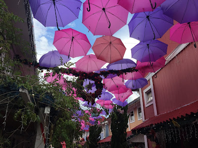 Entre os sobrados, guarda-chuvas abertos nas cores lilás, rosa e vermelho pendurados