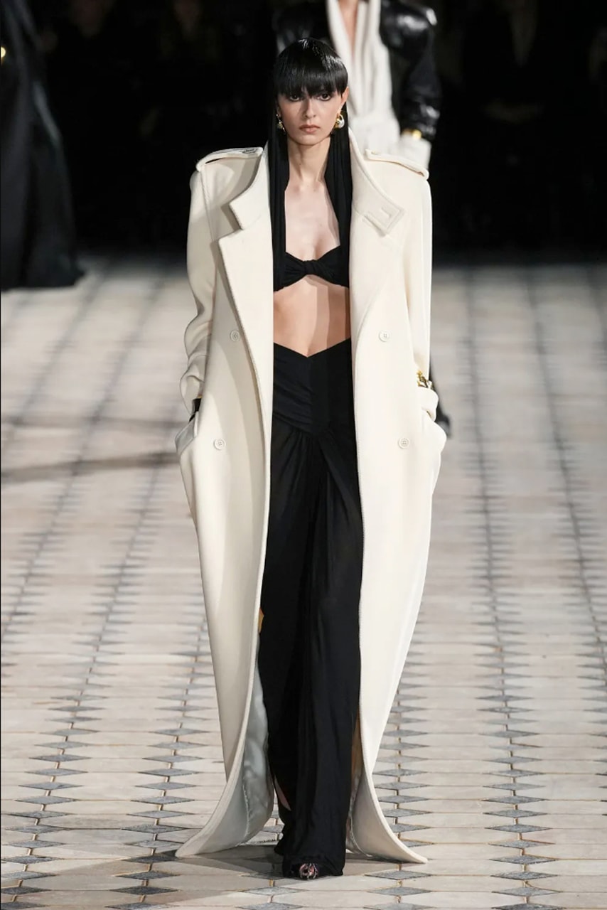 Woman with long straight black hair and blunt straight bangs and dark black eyeliner wearing chunky silver earrings, a cream white oversized trench coat, black palazzo trousers with a draping waist, walking with her hands in her pockets on a black and white tiled floor