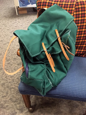 A dark hunter green knapsack with white stitching, a navy zipper running up the side, and pair of tan leather straps and silver buckles closing the top flap and outer pocket, sitting on a blue chair with a red, orange, and black plaid flannel pillow leaning against the back.