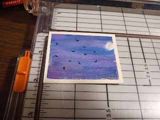 [Image Description] Small painting laying on a paper trimmer. Painting is of bats flying in the night sky with a full moon in the background.