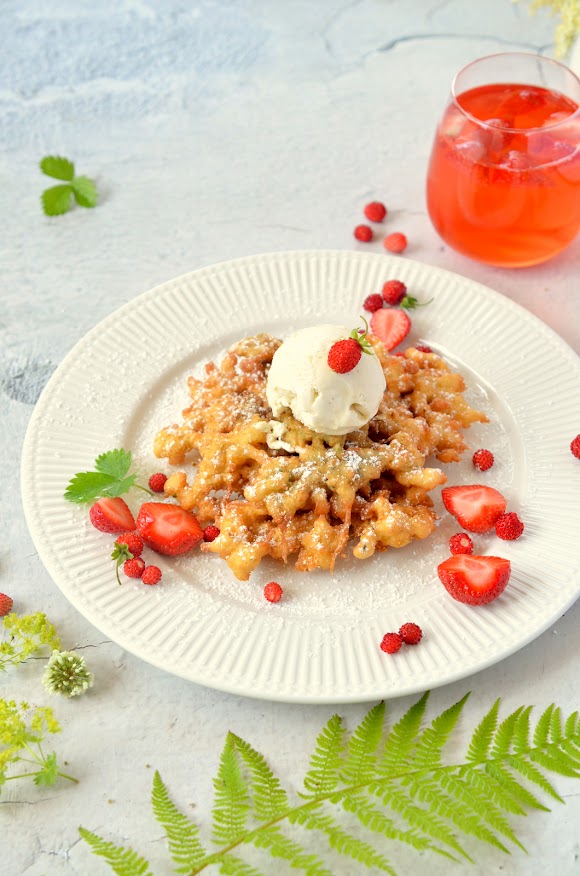 Blüten wurden in Backteig getaucht und ausgebacken – dieser Blütenkrapfen liegt auf einem weißen Teller, darauf eine Kugel Vanilleeis und rundherum frische Erdbeeren.