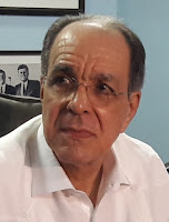 En la fotografía, Joaquín Ricardo está sentado de frente mirando hacia la izquierda. Pelo negro con amplias entradas. Rostro alargado, cejas pobladas con el ceño fruncido, gafas recetadas con montura muy delgada, tez clara, camisa blanca. Detrás, a su izquierda se encuentra una fotografía de Kennedy con otro acompañante.