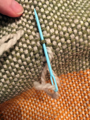 Close up of the same light green and white block, showing a marigold and white block below. The light blue plastic needle is inserted under a green thread while passing over the floats at either side of it. 
