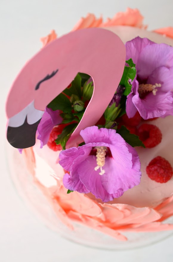 Eine Torte, die aussieht wie ein Flamingo – mit rosaroten Federn aus Schokolade und einem selbst gebastelten Kopf aus Karton. Die Torte ist mit lilafarbenen Hibiskusblüten und frischen Himbeeren dekoriert.