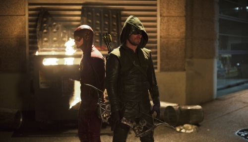 Grant Gustin as The Flash and Stephen Amell as The Arrow standing together in a street scene facing opposite directions