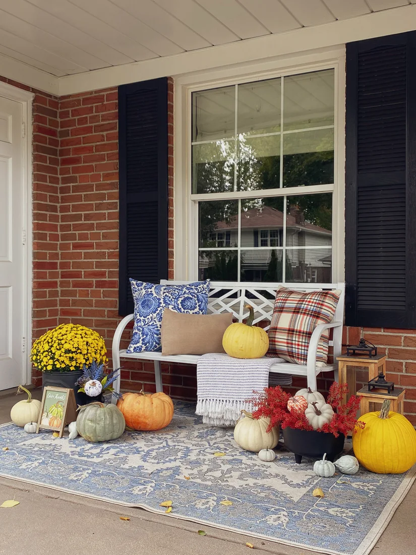 Fall front porch decor with white bench, plaid pillow, yellow mums, grey, white and orange pumpkins, and a blue and white traditional outdoor rug