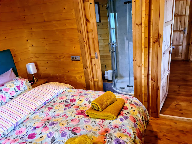 Image of interior of wooden lodge holiday accommodation. Double bed with floral bedding and teal headboard. Yellow towel bale on the bed and a view into the en suite shower room showing curved shower cubicle.