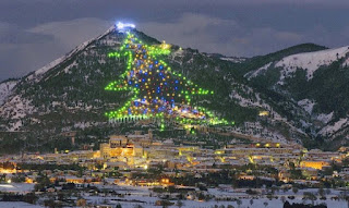 Nota: Gubbio é uma comuna italiana da região da Umbria, província de Perugia, com cerca de 30.453 habitantes.  Descrição: Foto noturna com vista panorâmica da cidade de Gubbio, na Itália. Ao longe, ao pé de uma colina bem alta, a cidade iluminada e nevada. Ao longo da colina com vegetação e neve, uma árvore de Natal composta por luzes verdes delineando o contorno do pinheiro e preenchido por outras luzes coloridas nas cores, amarelo, vermelho e azul, e bem no alto, uma luz branca em formato de estrela cadente. Com esta imagem, desejamos a todos amigos um Feliz Natal!