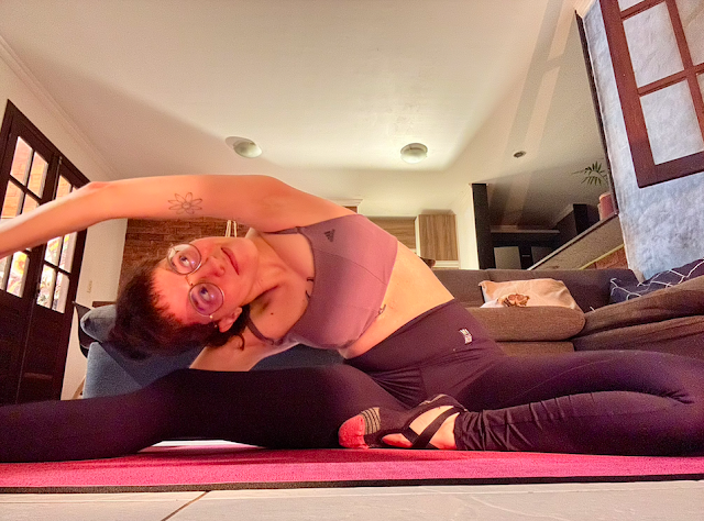 Foto da minha pessoa alongando para o lado esquerdo com o braço esticado tocando no pé com as pernas abertas em espacate no tapete de yoga