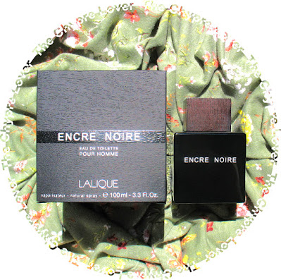 The black opaque bottle and black cardboard box of Encre Noire by Lalique (eau de toilette) lying flat on a flowered light green fabric