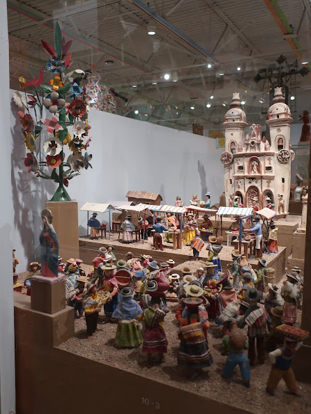 Photo of Mexican folk art in a display case. The display includes a toy cathedral, many dolls in colorful outfits standing facing the cathedral, some tents indicating a street market. A carved treelike structure with carved flowers and a carved statue of the Virgin Mary are also visible.