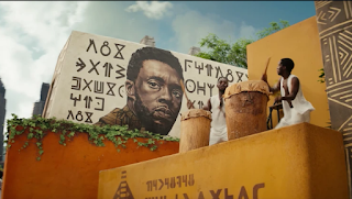 A picture of T'Challa painted on the side of the building with Wakandan writing around it. Two women are playing drums.