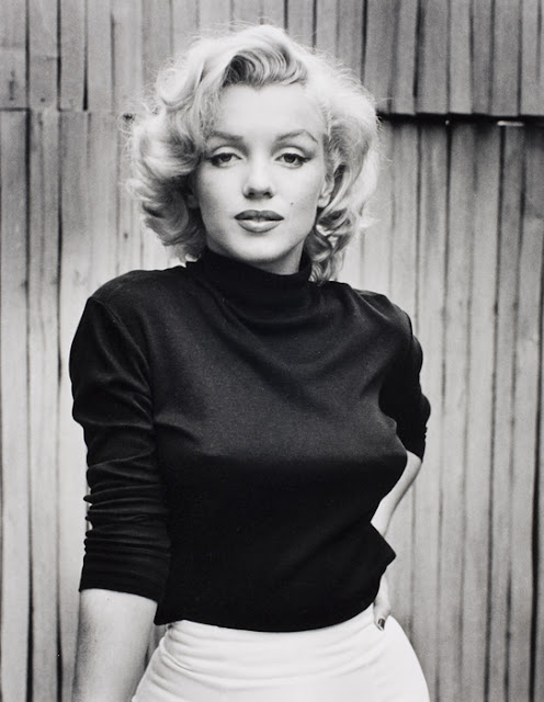 Foto da estrela de cinema Marilyn Monroe em preto e branco. Marilyn olha para a câmera, seu braço esquerdo está apoiado na cintura e a mão direita está apoiada em um objeto que não aparece. Ao fundo vemos uma cerca de madeira.