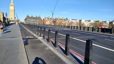 A view along Westminster bridge. The houses of Parliament are in the distance. On the bridge, there is a wide footway to the left, then a cycle track to its right and then a skinny island with bollards. There is the a bus lane.