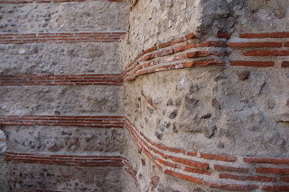 A photo of a Roman wall with alternating bands of stone/cement with thin red brick,. The wall has three segments. One segment starts at the left, then there's a corner and the short second segment is perpendicular to the first. A second corner close to the viewpoint, and the third segment is parallel to the first and runs off the right edge of the photo.