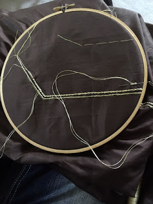 Smooth brown silk stretched in a round wooden embroidery hoop, with bright green basted outlines and a fine gold split stitch just inside the outlines. The beginning of a line of chain stitch follows the split stitch, and a needle threaded with doubled gold thread has just made two chain stitches about half an inch inside the first chain stitch line.