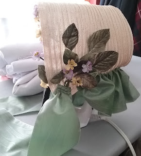 Left side view of a low-brim natural straw bonnet, with green curtain; along the neckline there is a large cluster pale green leaves with a few yellow and purple flowers.