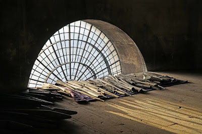 A row of damaged organ pipes on a wooden floor, in front of an arched window with many small panes.