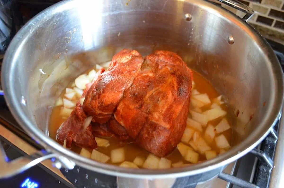 A pork shoulder roast in a stainless steel pot rubbed with herbs and spice. Garlic Onion, and Citrus Juice are added to the pot.