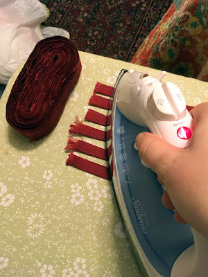 An oval disk of red linen strips on the end of an ironing board, next to the ends of six strips being pressed in half lengthwise with a blue-and-white iron, on a pale green floral ironing board. A red oriental rug and the edge of a pink, orange, yellow, and turquoise patchwork patterned bedspread fill the background.