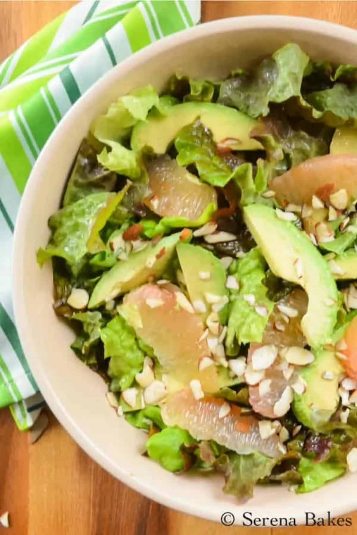 A down shot of Grapefruit Avocado Salad in a cream colored bowl on a wooden cutting board with a white and green stripped kitchen towel in the top left corner under the bowl..