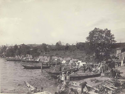 black & white photo - about 5 long canoes pulled onto shore, with men and women occupied on shore; building in background