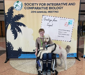 Amy-Charlotte Devitz and service dog Fisher in front of a “Society for Integrative and Comparative Biology" sign.