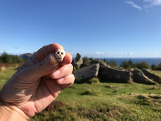 A photo of a small, ceramic skull (Skulferatu #85) being held up with the Giants' Graves in the background.  Photo by Kevin Nosferatu for the Skulferatu Project.