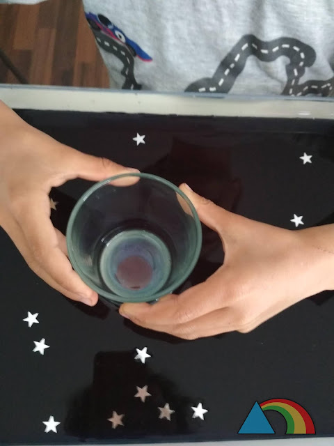 Niño buscando planetas en una bandeja transparente con agua negra, con la ayuda de un vaso