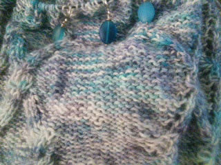 a column of reverse stocking stitch, with cabled lace on either side.  It's knit in a variegated blue-grey fingering weight yarn,  and three sea glass stitch markers are on the circular needle cord at the top of the work