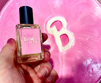A white hand holding a rectangular glass bottle with a cyldienxjal black lid with a pink square label that says barbie x lush in white font on a bright background