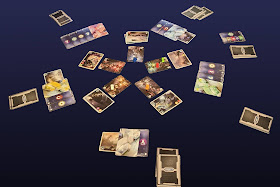 A game of Multiuniversum in progress. The five generator cards are arranged in a five-spoke circle in the centre, the scientist meeples on them, with the stacks of portal cards just outside of the circle corresponding to each generator card. The players' lab cards are around those; some of them have tool cards and/or portal cards tucked under the lab cards. A stack of cards representing each player's hand is near that player's lab card. The deck of action cards and the discard pile are off to one side.