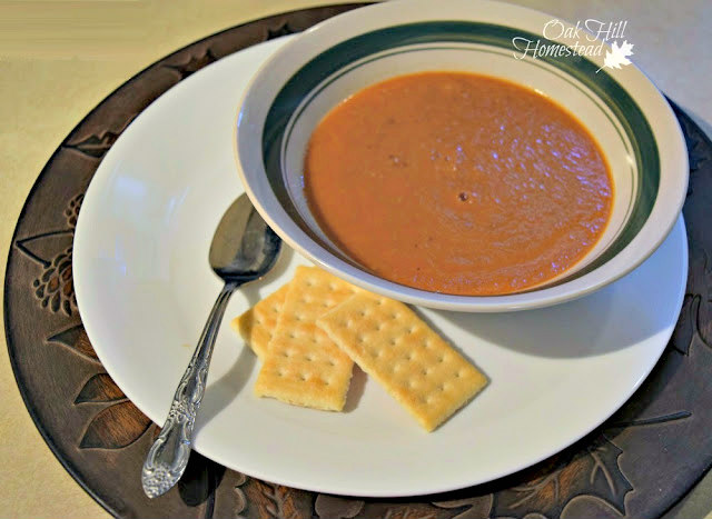 A bowl of tomato soup on a white plate and a brown charger, with spoon and crackers.