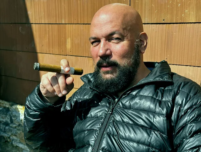 12/12 bald man wearing a puffy leather jacket sitting outside smoking a cigar