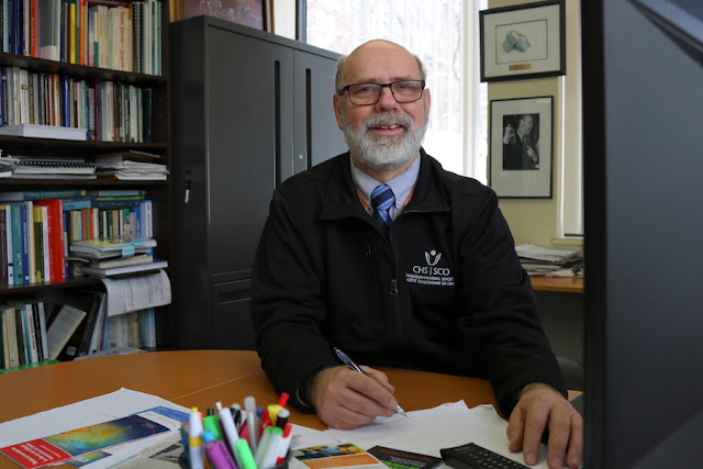 Gary Malkowski en un despacho con un bolígrafo en mano ante una mesa, libros al fondo y sonriendo