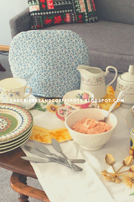 A blue and white handmade tea cozy with a spread of afternoon tea