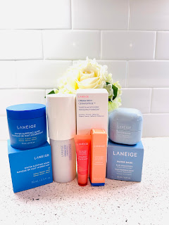 A beautifully arranged lineup of Laneige skincare products sits atop a pristine white surface. From left to right, the products include Laneige Lip Glowy Balm, Water Bank Blue Hyaluronic Cream Moisturizer, Water Sleeping Mask, and Cream Skin Toner Moisturizer. Each product is elegantly packaged in sleek containers, showcasing the brand's commitment to both style and effectiveness. The vibrant colors and clean design of the packaging invite the viewer to embark on a journey to glowing, radiant skin with Laneige.