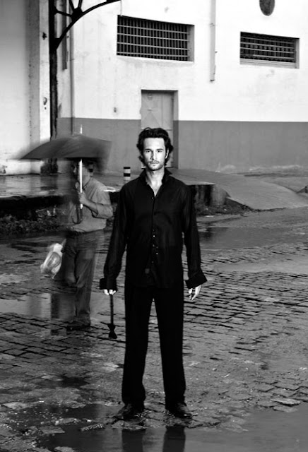 Retrato do ator Rodrigo Santoro feito pelo fotógrafo Jorge Bispo. Na foto, o ator é visto de corpo inteiro e em pé em uma rua a céu aberto. Usa traje social na cor preta e encara a câmera com o corpo estático. O chão é irregular e há poças de água. Logo atrás do ator, um homem de guarda-chuva passa com um borrões indicando movimento.