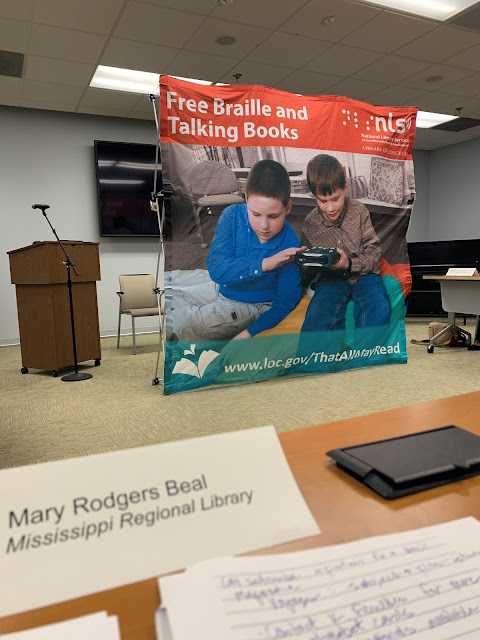 Sitting next to a lectern and microphone is a large banner that says free braille and talking books and has a picture of two boys using a TBS machine. A paper nameplate that says Mary Rodgers Beal is in the foreground.