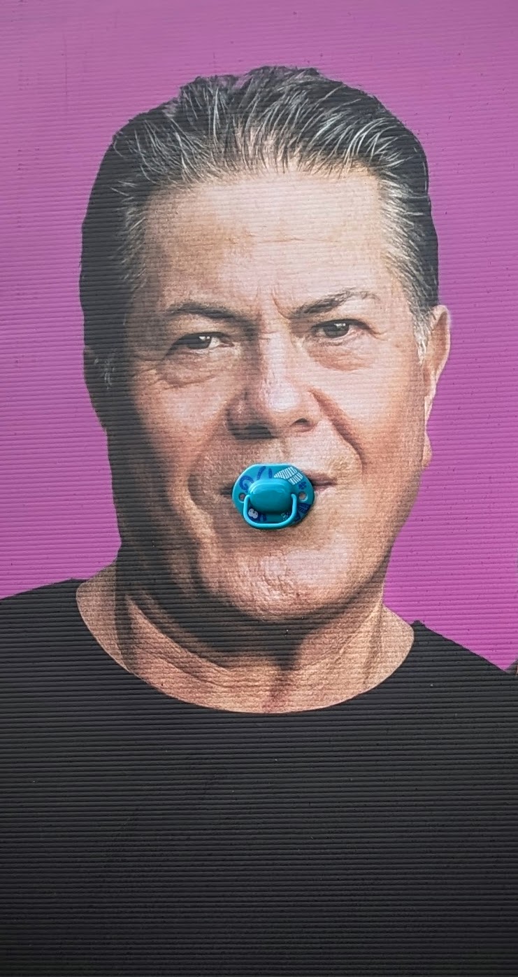 Election poster photograph of Brian Tamaki, head and shoulders shot against a pink background. A real blue babies dummy has been placed into his poster mouth.