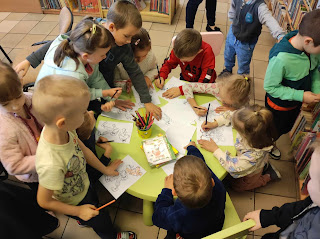 Sala biblioteczna. Grupa dzieci otacza zielony niski plastikowy stolik, część dzieci stoi a część siedzi. Na stoliku stoi kubek z kolorowymi kredkami i kolorowanki, które dzieci kolorują.