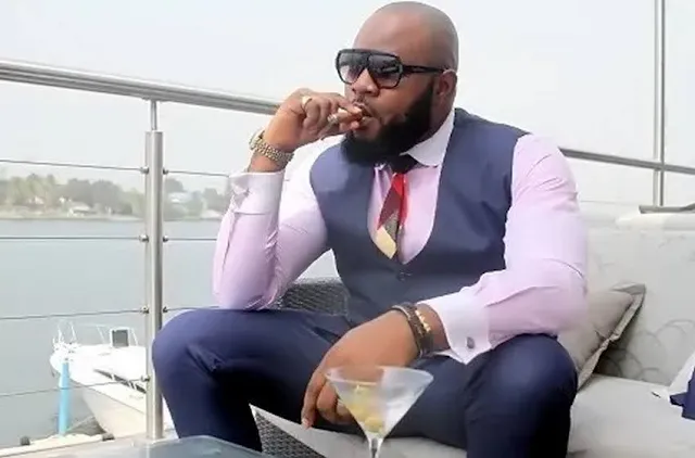 Bald black man with sunglasses on and a suit smoking a cigar on a boat with a city in the background